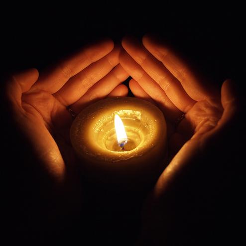 Hände halten eine brennende Kerze in der Dunkelheit, erzeugen warmes Licht und Schatten.